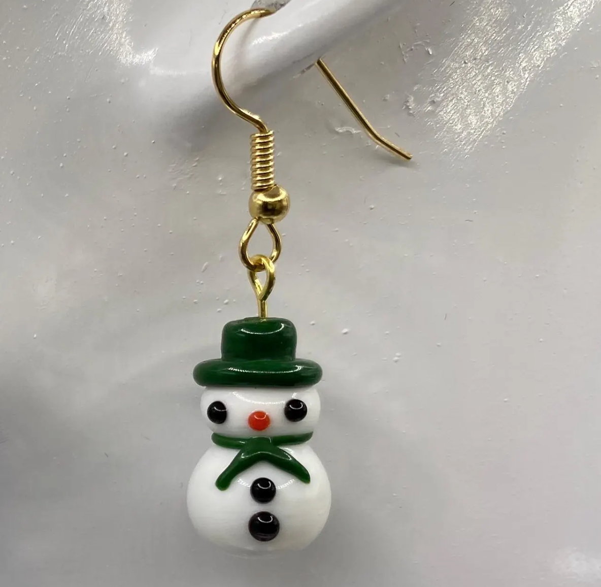 white lampwork snowman earring with green hat and scarf hanging from mannikin's earlobe