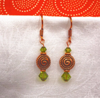 A pair of handmade copper drop earrings with spiral charms and green faceted crystal beads, displayed on a white textured fabric background with a red patterned fabric at the top.