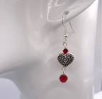 A single dangling earring displayed on a white mannequin ear. The earring features a silver metal hook, a small red faceted crystal bead, an ornate silver heart charm with scrollwork, and a larger red faceted crystal bead dangling below.
