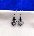 A pair of handmade dangle earrings featuring white and black bumpy beads, black faceted crystal beads, and silver fishhook wires, displayed on a white textured surface with a vibrant blue background.