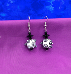 A pair of dangling earrings with white lampwork beads featuring black and white bumps, black crystal beads, and silver fishhook ear wires, displayed on a purple textured surface against a blue background.