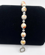 A beaded bracelet displayed on a black velvet jewelry stand. The bracelet features glossy white beads with orange and blue floral patterns, separated by small white faux pearls and silver-toned spacer beads, ending in a silver-toned circular clasp.