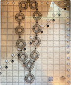 A necklace of gold plated brass wire wrapped rings on a clear acrylic quilting ruler with a grid and measurement markings, laid flat on a light wood surface. 