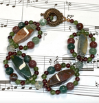 A beaded bracelet with polished gemstone beads in green, burgundy, and gray, featuring large natural stone rectangles encircled by smaller jasper rounds, which fastens by a copper circular clasp, displayed on a sheet of printed music.