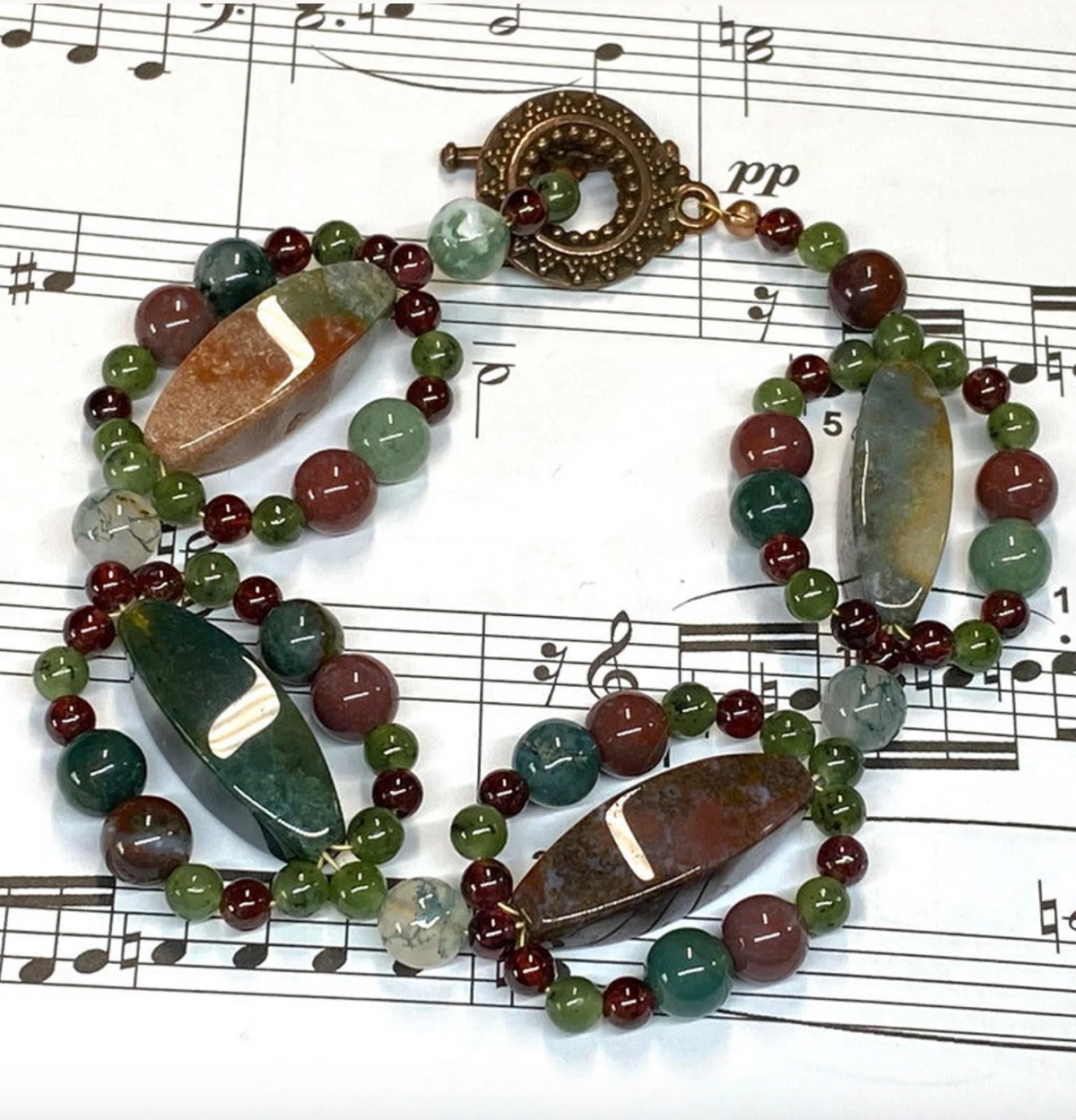 A beaded bracelet with polished gemstone beads in green, burgundy, and gray, featuring large natural stone rectangles encircled by smaller jasper rounds, which fastens by a copper circular clasp, displayed on a sheet of printed music.