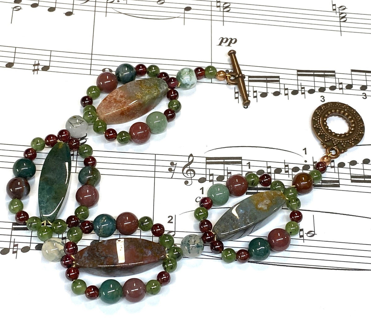 A beaded bracelet with natural stone beads in green, red, brown, and gray, featuring a copper toggle clasp, laid out on a sheet of musical notation.