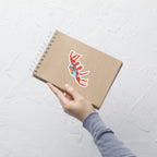 Hand holding a brown notebook with a red and blue crab sticker on a light gray textured background