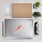 Red and blue crab sticker on a laptop on a desk with a notebook, mouse, pencil, and glasses.