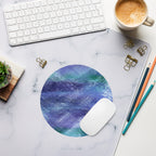 A round mouse pad with a blue and purple watercolor texture, styled on a white marble desk mat with a white computer mouse, keyboard, coffee cup, and office accessories.