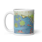 A white ceramic mug featuring a whimsical, hand-painted style illustration of a pond scene with a yellow and black butterfly, green lily pads, red tadpoles, and reddish pink and green frogs.