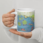 A white ceramic mug with a colorful painted nature scene of a pond, lily pads, a butterfly, and small small and large frogs, held by two hands against a light gray background.