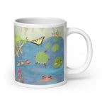 A white ceramic mug featuring a colorful printed illustration of a pond scene with frogs, lily pads, a butterfly, and a large frog on a white background.